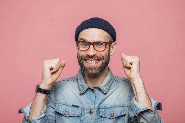 Mavi gözleri ve neşeli ifade ile mutlu sakallı erkek yumruk clenches, kazanan olmak nasıl bir duygu, positiveness, pembe bir arka plan üzerinde izole gösterir. Heyecanlı duygusal genç adam kapalı poz veriyor
