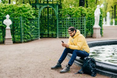 Sakallı hippi adam açık atış şık çeşme sörf sosyal ağlar üzerinde yeşil park arka plan izole cep telefonunu kullanarak Parkı dinlenme giyinmiş. Ciddi adam yana doğru çekim