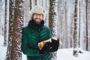 Gülümseyen erkek giyer sıcak kış giysileri tutar yakacak odun yakışıklı, standları tree, yakınındaki kış ormanda piknik olacak arkadaşlarınızla boş zaman harcıyor, soğuk havalarda sıcak yapar ateş