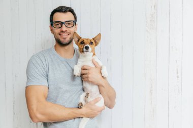 Çekici neşeli Kafkas erkek casual t shirt, gözlükler, favori Evcil hayvan tutar, Beyaz ahşap arka plan boşluk ile karşı duruyor. Onun mutlu tıraşsız adamla jack russell terrier