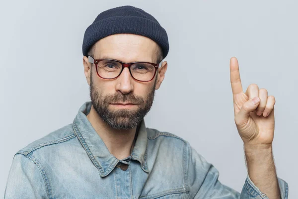 Serious bearded man wears glasses, fashionable hat and denim jacket, raises fore finger as tries to warn you about something, isolated over grey background. Just hear me, please! Studio shot