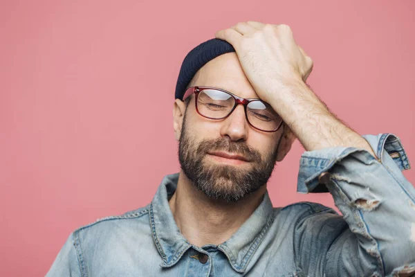 Portrait of depressed middle aged unshaven male closes eyes and keeps hand on forehead, has unhappy expression, isolated over pink background. Fatigue hipster guy has headache after hard work