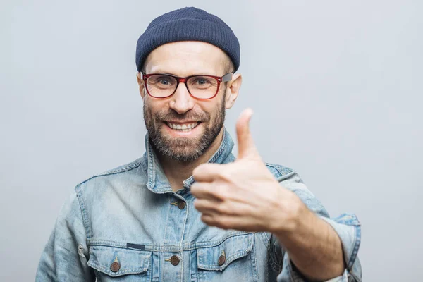 Happy delighted man with stubble raises thumb as shows his approval, dressed in stylish clothing, isolated over white studio background. Satisfied middle aged male gestures indoor. Body language
