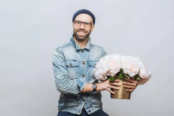 Happy delighted male buys bouquet of flowers for special occasion, wears denim jacket and denim shirt, isolated over white background. Joyful emotional man with present. Beautiful flowers for you