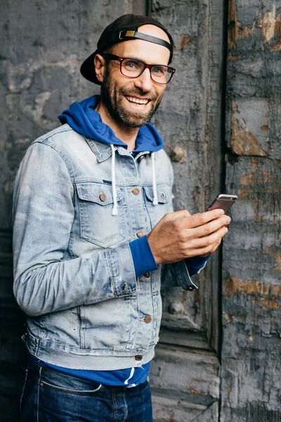 Stunning man with blue eyes, beard and broad smile wearing cap, denim shirt and eyeglasses holding smartphone isolated over old wall background. Pleased man in stylish clothes using cell phone