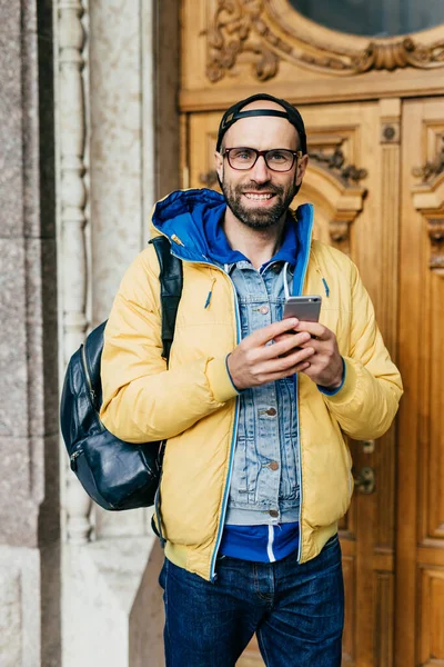 Young traveler in trendy clothes standing in museum having happy look to be there. Cheerful bearded male in yellow jacket holding rucksack on his back holding mobile visiting regional museum