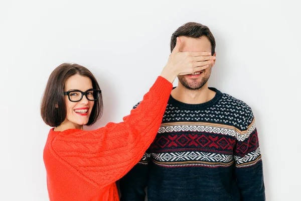 Portrait of happy covers eyes to her husband, prepares surprise for him, wears casual red oversized sweater, has positive smile, isolated over white background. I am going to show something to you!