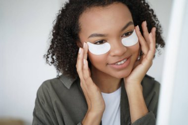 Happy beautiful african american girl applying moisturizing cotton under eye patches to reduce puffiness, remove dark circles. Beauty treatment for young skin, skincare cosmetics advertising.