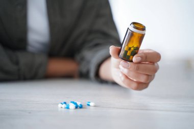 Close up of female patient holding jar with pills, vitamins, medication capsules for cure and wellness. Woman considering daily dose of dietary supplements or medicine remedy, sitting at table.