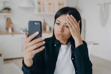 Modern young woman blogger holding smartphone, makes video call or takes a selfie photo, standing at home. Focused female has online communication on cellphone, using mobile phone camera.