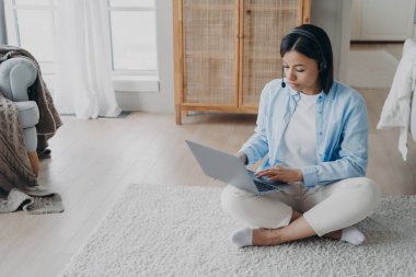 Focused female freelancer wearing headset works on laptop, sitting on the floor at home. Serious woman in wireless headphones watching educational webinar, studying online. E-learning, remote job.