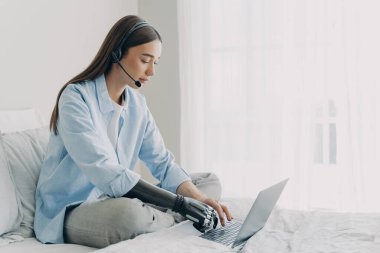 Handicapped freelancer has online meeting sitting on her bed. Young caucasian woman with artificial arm is working on laptop in bedroom. Girl is using headphones and microphone and chatting.