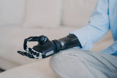 Cyber hand of disabled person which is sitting in lotus pose on couch. Myoelectric bionic prosthesis. Artificial limb with nervous sensor control. Robotic arm and hand in meditation.