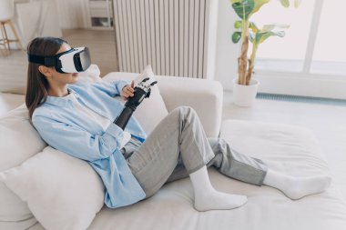 Attractive disabled girl is relaxing in vr glasses on couch. Handicapped caucasian person gets rehabilitation for sensor cyber arm. Amputee is setting functions of myoelectric robotic limb.