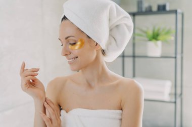 Mutlu kız altın göz bantları yapıştırıyor ve ellerinin ipeksi tenine dokunuyor. Spa ameliyatlarından sonra havluya sarılmış çekici Avrupalı kadın. Genç İspanyol bayan evde saç ve vücut bakımı yapıyor..