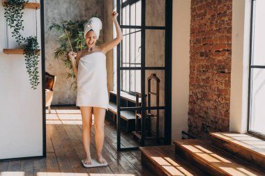 Weight loss concept. Excited slim european woman taking step onto scale to measure her weight. Girl wrapped in towel after bathing has time at spa. Overjoyed woman checking weight after shower.