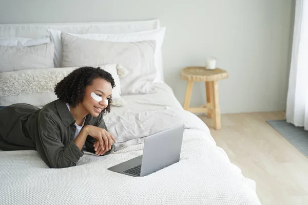 Mixed race young girl with under eye patches on face watching series on laptop lying on bed at home. Smiling pretty lady with afro hair shopping online while skincare daily routine in bedroom.