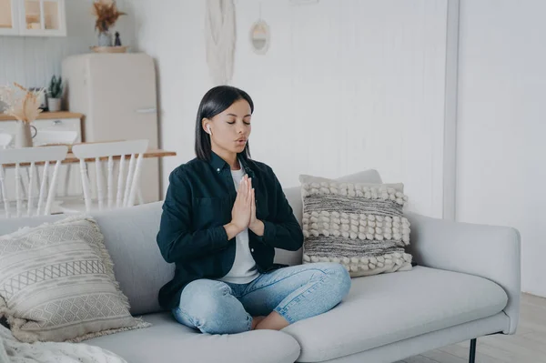 Calm young female deep breathing, practicing yoga, sitting in lotus pose on couch at home. Woman meditates folded hands in namaste gesture, relaxing on sofa. Healthy lifestyle, stress relief concept.