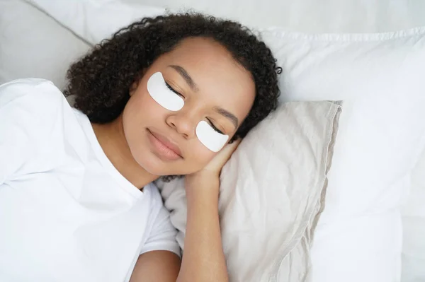 Young biracial girl with under eye patches on face relaxing lying in bed with closed eyes. Pretty woman using undereye skin beauty masks, preventing bags, reducing wrinkles. Morning skincare routine.