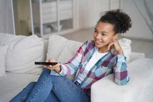 Teenage girl enjoying tv series watching and shifts channels with remote controller. Afro young woman has leisure in morning and sitting on couch on quarantine. Reality show watching.