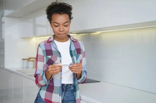 Unwanted pregnancy. Teenage girl gets in trouble with positive pregnancy test result. Frustrated african american young woman is holding stripe test and looking at it. Contraception concept.