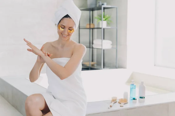 Lady applies eye patches and moisturizing skin with lotion. Attractive woman wrapped in towel after bathing is sitting on bathtub. Relaxation at spa resort. Modern interior of bathroom.