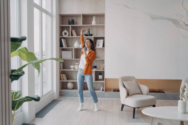 Oturma odasında deli gibi eğlenen bir kız. Mutlu İspanyol kadın mikrofondaki gibi evde telefonla şarkı söylüyor. Rahatlama ve dans. Duygusal ifade ve isyan kavramı.