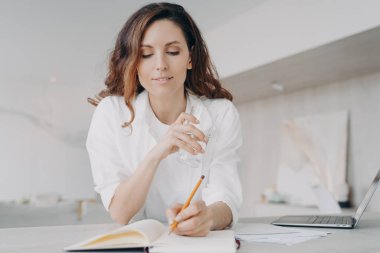 Beyazlı genç Avrupalı kadın notlar alıyor. Serbest çalışan ya da iş kadını programa bakıyor. Mutlu kız masadaki deftere bir şeyler yazıyor. Karantinada evden uzak çalışma.