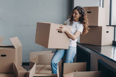 Beyaz tişörtlü genç bayan karton kutu taşıyor. Gülümseyen çekici İspanyol kadın eşyalarını toplayıp gidiyor. Paketlenmiş kutular yerde. Taşınma ve yeni apartman konseptine geçiş.