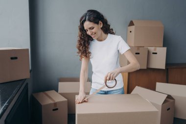 Young spanish woman is packing boxes with duct tape. Moving service business. Lady is wrapping cardboard boxes with packing tape. Female worker preparing boxes for shipping and storage.