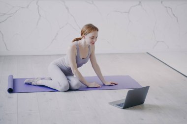 Young caucasian woman practicing yoga on mat on floor of her room. Internet fitness lesson with computer on self isolation. Concept of e-learning and home online classes on corona virus quarantine.
