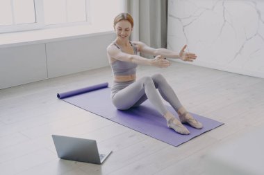 Slim sporty caucasian girl in sportswear is stretching on mat at home. Active healthy lifestyle. Concept of distance learning and online fitness classes. Personal training on coronavirus quarantine.