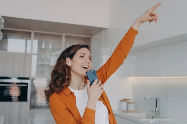 Favourite song karaoke singing at home. Happy caucasian woman in airpods is singing with mobile telephone as with microphone. Girl having fun, relaxing and dancing. Online music listening.