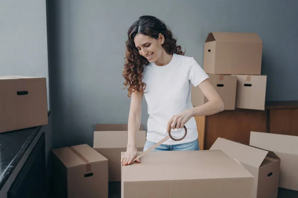 Young spanish woman is packing boxes with duct tape. Moving service business. Lady is wrapping cardboard boxes with packing tape. Female worker preparing boxes for shipping and storage.