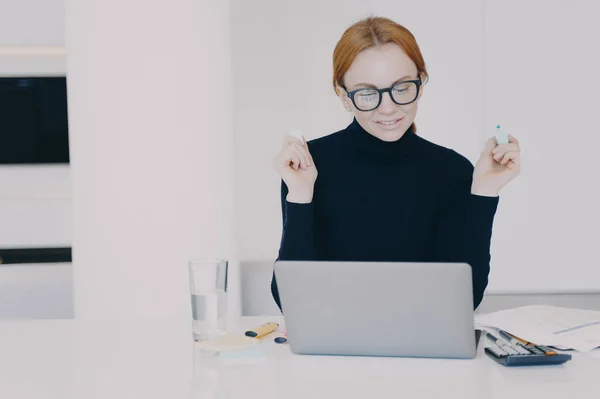 Confident female entrepreneur at work. Red haired woman in office is doing paperwork with marker pen and highlighting text. Positive caucasian girl entrepreneur is working on project at laptop.