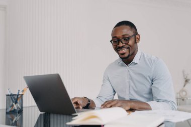 Düşünceli Afro-Amerikalı gözlüklü adam internette dizüstü bilgisayarla çalışıyor, ofis masasında oturuyor, iş projesinden memnun. Gülümseyen zenci eleman işyerindeki işinden memnun..
