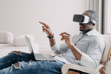 Relaxed businessman clicks virtual buttons. Gestures of african american man in vr glasses. Guy is working at home. Distant work and futuristic gaming. Modern digital technology for business.