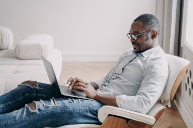 Online freelance job. Handsome african american man in front of pc. Relaxed happy businessman in formal wear is sitting in cosy chair at his apartment. Startup project, remote work.