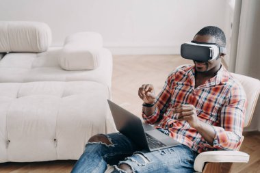 Afro man in vr headset. Workplace at home office. Businessman has virtual meeting. Relaxed freelancer is sitting in chair and working on project. High tech digital technology and vr gaming.