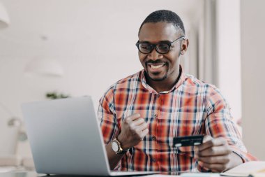 E-banking, e-commerce. African american man use e banking service at laptop. Black guy satisfied by easy secure payment, receiving benefits as loyal client, paying online, holding plastics credit card