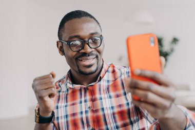 African american man wearing glasses holding smartphone having video call conversation, gesturing, takes selfie. Happy black guy looking at mobile phone smiling reading good news message.