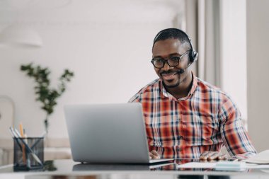 African male student wearing headset distance learning language online at laptop sitting at desk at home. Black guy wears headphone watching webinar, listening lecture. E-learning, remote education.