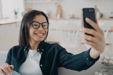 Video görüşmesi, uzaktan konuşma. Mutlu melez kadın rahatlıyor ve akıllı telefonda sohbet ediyor. Beyaz kadın evde eğleniyor ve gülümsüyor. Aygıt ve uygulama kullanarak.