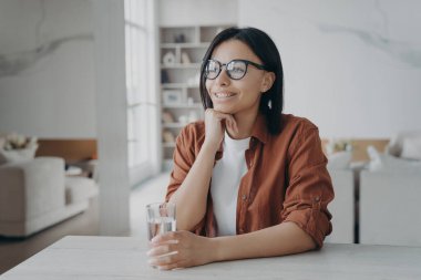 Smiling young woman enjoy purified mineral water holding glass sitting at kitchen at home. Happy female wearing glasses with pure water drink at table. Healthy lifestyle, diet concept.