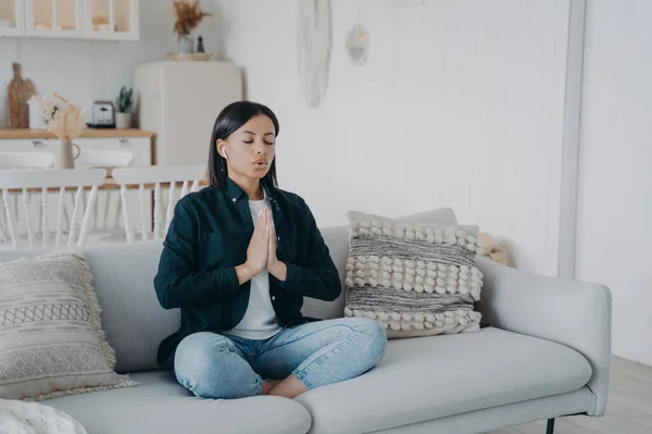 Calm young woman deep breathing practicing yoga sitting on couch at home. Female meditates folded hands in namaste gesture in lotus posture on sofa. Wellness, stress relief concept.
