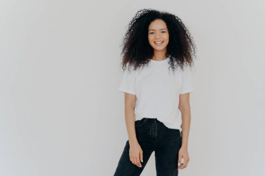 Kıvırcık saçlı, ince vücutlu, beyaz tişörtlü ve siyah kotlu gülümseyen bir kadının fotoğrafı, mutlu yüz ifadesi, beyaz arka planda modeller, reklam içeriğiniz için kopyalama alanı