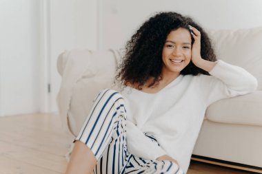 Koyu tenli, güzel bir kadın, kanepenin yanında oturuyor, beyaz kazak ve çizgili pantolon giyiyor, olumlu duygulardan hoşlanıyor, hafta sonunu evde geçiriyor. İnsanlar, dinlenme, mutluluk kavramı