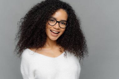 Indoor shot of happy young lady has Afro hairstyle, smiles broadly, glad to be promoted, wears optical glasses and white sweater, models over grey background. Ethnicity, emotions, pleasure concept