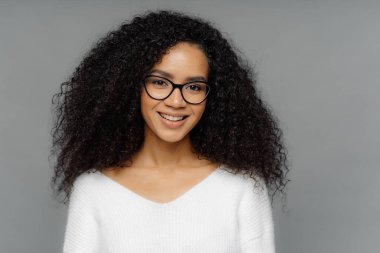 Afro saç modeli ile Muhteşem güzel kıvırcık kadın, mutlu hissediyor, kamera hafifçe gülümsüyor, optik gözlük ve beyaz kazak giyer, gri arka plan üzerinde izole. Mutlu duygular ve duygular kavramı