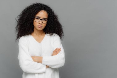 Kendine güvenen koyu tenli etnik kadın elleri göğüs üzerinde çapraz tutar, kamera ciddi bakar, optik gözlük ve beyaz kazak giyer, boş alan ile gri arka plan karşı duruyor